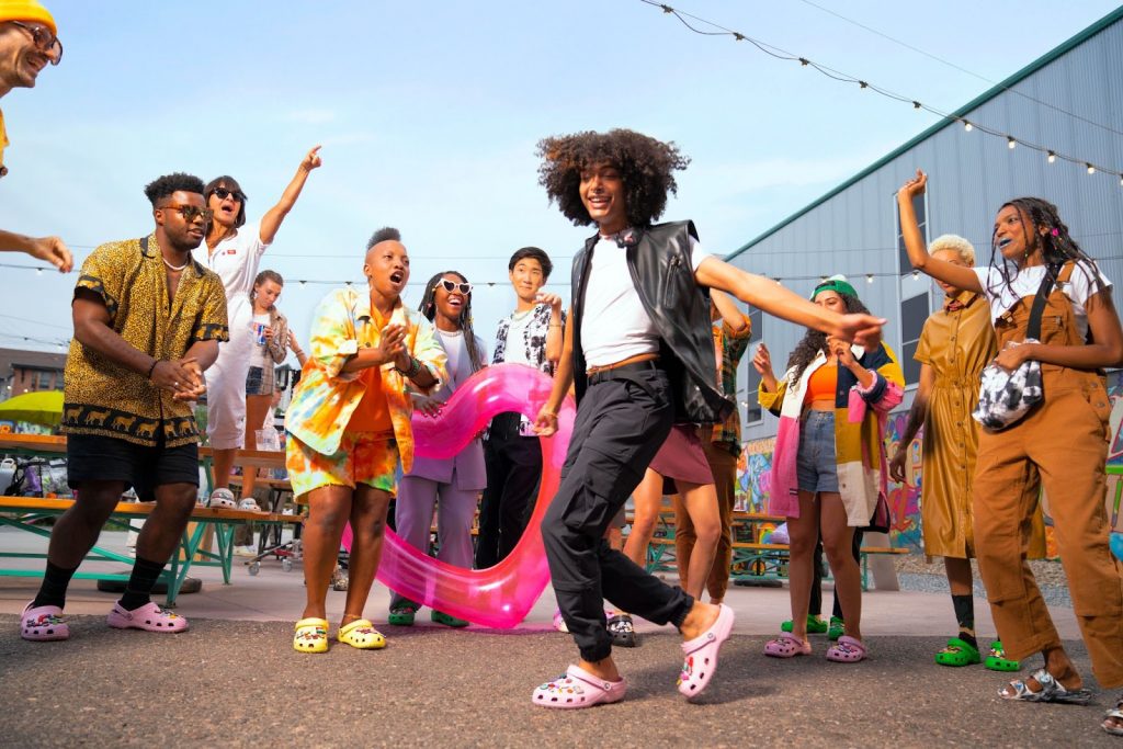 Portal Jaciara Barros – Um grupo comemora 20 anos ao ar livre enquanto uma pessoa sorridente, de Crocs rosas e roupas preto e branco, dança. – Portal Jaciara Barros