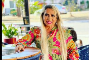 Portal Jaciara Barros – Gilda Bonfim, de cabelos loiros e vestido floral, sorri ao lado de uma mesa redonda durante celebração de aniversário. – Portal Jaciara Barros