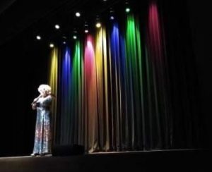 Portal Jaciara Barros – Pessoa com vestido brilhante está no palco do Teatro Feevale, com luzes coloridas iluminando cortinas fechadas. – Portal Jaciara Barros