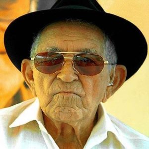 Portal Jaciara Barros – Idoso nordestino com óculos escuros, chapéu preto e camisa branca posa sério durante o Dia do Nordestino. – Portal Jaciara Barros