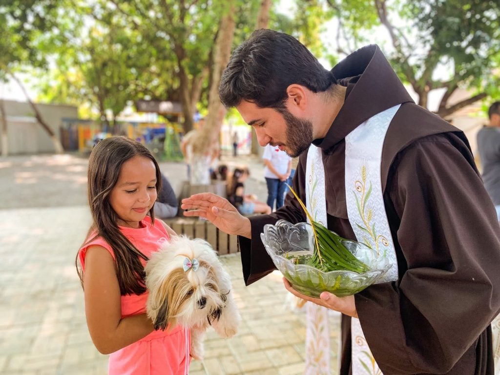 Portal Jaciara Barros – Um homem de túnica marrom realiza a bênção dos animais, abençoando um cachorro branco no colo de uma menina ao ar livre. – Portal Jaciara Barros