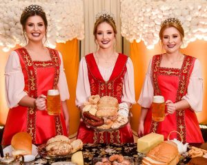 Portal Jaciara Barros – Três mulheres com vestidos tradicionais vermelhos participam da 17ª Kerbfest com canecas de chope e mesa de comidas variadas. – Portal Jaciara Barros