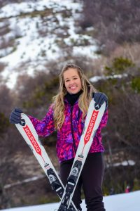 Portal Jaciara Barros – Thayná Costa posa na neve com jaqueta rosa de inverno, segurando esquis em pé, com árvores e colinas ao fundo. – Portal Jaciara Barros