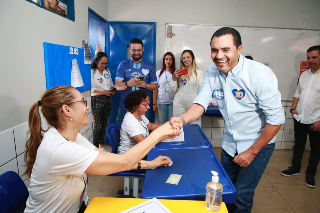 Portal Jaciara Barros – Homem sorri e cumprimenta mulher sentada em seção eleitoral de Taquaruçu, enquanto outras pessoas observam e registram o momento. – Portal Jaciara Barros